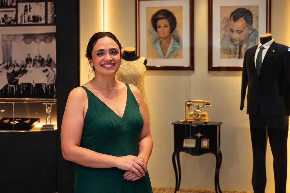 Vanessa Resende, diretora da MuseoLógica, em uma sala de exposição com retratos, vestido e terno em vitrine